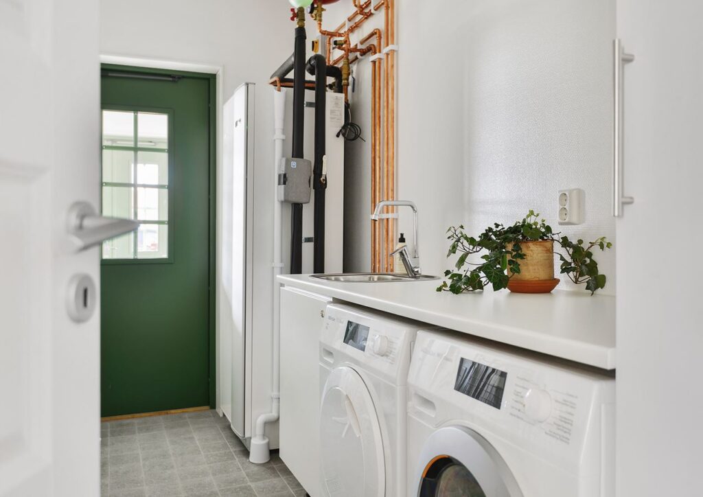En bild som visar en kombinerad groventré och tvättstuga. Grön ytterdörr med spröjsade fönster i bakgrund, värmepump, torktumlare och tvättmaskin i förgrund.